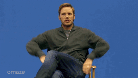 Celebrity gif. Chris Pratt sits atop a director's chair in front of a blue screen, which transforms into a wondrous, cosmic background as he theatrically raises his arms for us to behold the galaxy behind him.