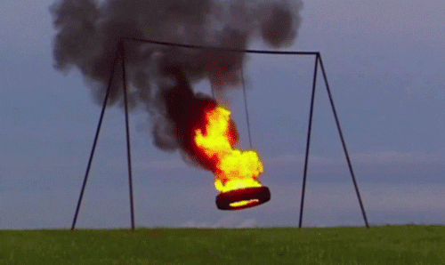 Video gif. Atop a green hill, a tire swing is on fire, burning with bright flames and releasing a swarm of black smoke into the indigo sky.