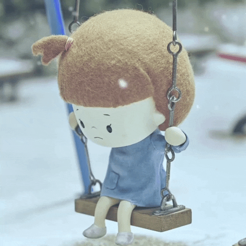 Stop-motion gif. Girl with brown hair and a tiny topknot sits on a swing, looking sad, as snow falls around her.