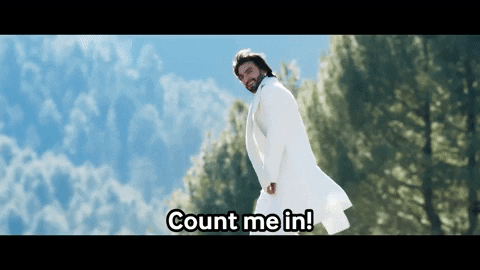 Movie gif. Ranveer Singh as Rocky in "Rocky Aur Rani Ki Prem Kahani" wears a white jacket and white scarf as he smiles and confidently walks across a forest background, shoulder-length hair blowing in the breeze and looking suave. Text, "Count me in!'