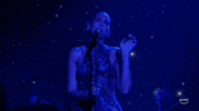 ACM Awards gif. While gripping mic stand with one hand Dua Lipa waves free hand at ear-level height before bringing it down then back up again as she sings and lightly shakes her head from side-to-side.