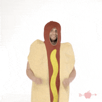 Video gif. A man in a giant hot dog costume raises his arms and crosses his fingers like he's wishing for something.