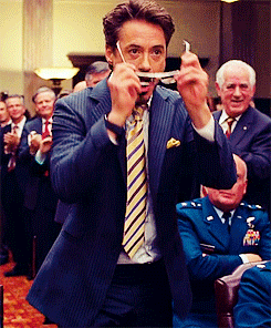 Movie gif. Robert Downey Jr as Tony Stark in Iron Man slides on sunglasses with a smooth grin on his face. He gives a thumbs up as people in business attire and military uniforms stand and clap behind him. 