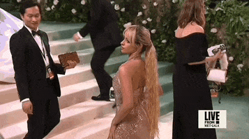 Met Gala 2024 gif. Camila Cabello poses to show the back of her Ludovic de Saint Sernin shimmery gold halter gown with a deep v-neckline. Her hair is blonde with brown roots and styled in a long wavy ponytail that falls to her buttocks. She's holding a purse that is an actual rectangular block of ice with a rose inside