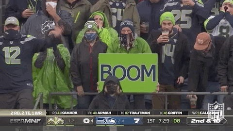 Sports gif. We zoom in on NFL Seahawks fans standing in the audience at a game wearing raincoats and beanies, looking cold but happy. One man displays a large sign that says, "Boom."