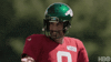 Sports gif. Coach Aaron Rodgers in a red jersey and green Jets helmet gives someone offscreen a cheesy smile as he raises a finger to point at them. 