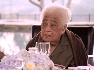 Video gif. Elderly woman sits at a flower topped dining table by a pool and stares straight-faced at someone before letting out a disappointed sigh and shaking her head.