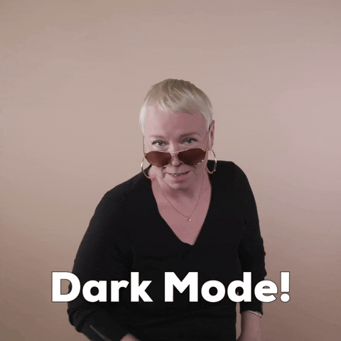 Reaction gif. A white woman with invisible disabilities, with hair styled in a sleek platinum pixie cut and big hoop earrings slide her aviator sunglasses up the bridge of her nose, gives two thumbs up and declares, "Dark mode."