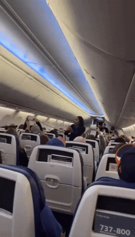 Video gif. Two rows on an airplane are playing a game against each other to see who can unroll a toilet paper the fastest by passing it to each person down the row. A flight attendant is in the middle cheering them on.