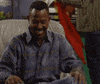 TV gif. Seated in a gray leather armchair next to Halloween decorations, a giddy Martin Lawrence from Martin laughs while holding his stomach and bouncing his knees.