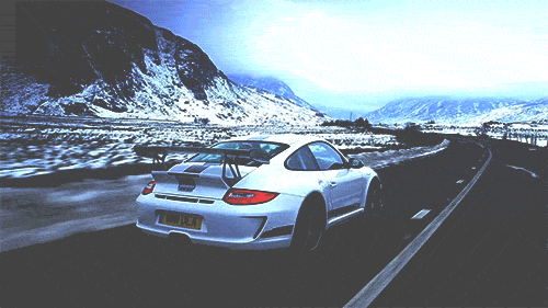 Video gif. White Porsche GT3 RS travels along a highway weaving through a vast, snowy mountain terrain.