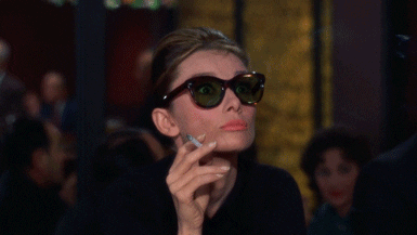 Movie gif. Aubrey Hepburn as Holly Golightly in Breakfast at Tiffany’s wears sunglasses and holds a smoking cigarette in between her fingers. Her eyes widen behind her tinted lens and her eyebrow raised in shock. She slides her sunglasses down her nose and looks over her glasses up at someone.