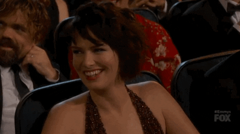 Video gif. Lena Headey at the 2015 Emmys glances over and smirks as if saying oops. 