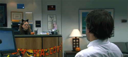 The Office gif. Jenna Fischer as Pam is dressed like a cat for Halloween as she sits at the Dunder Mifflin reception desk. John Krasinski as Jim sits across the room in the foreground without a costume. The two of them give each other a long-distance air-high-five.