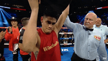 Sports gif. Slow motion clip of Boxer Emanuel Navarrete putting on a cowboy hat in a dramatic way as the referee holds one arm up and a man in the background passes by with a boxing belt.