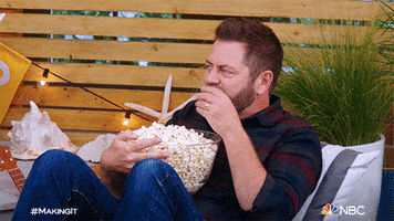 Celebrity gif. Nick Offerman, seated, stuffs his face with popcorn from a bowl. He shimmies around happily.