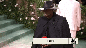 Met Gala 2024 gif. Usher, wearing an all-black custom Alexander McQueen ensemble of a cape, leather fedora, and red paper flower.