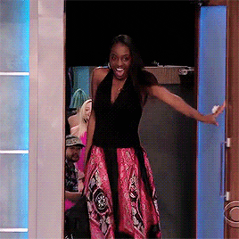 Reality TV gif. A beautiful woman with long, flowing hair in a black and red flowing dress steps onto a runway through a stage door and makes a dramatic, beaming entrance. 