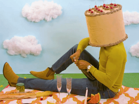 Video gif. Man with a cake for a head sits on picnic blanket and opens a bottle of sparkling wine, which foams and spills out the top. Text, "Congrats!"