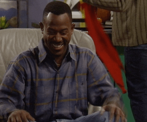 TV gif. Seated in a gray leather armchair next to Halloween decorations, a giddy Martin Lawrence from Martin laughs while holding his stomach and bouncing his knees.