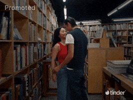 Sponsored gif. A man whisks a woman off the floor in a bookstore and spins her around. She wraps her arms around his shoulders, beaming with joy. The lights in the bookstore flicker as the camera draws back dramatically. Animated fireworks with stars and dotted lines move playfully. The Tinder logo is in the bottom right corner.
