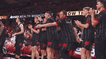 Sports gif. Members of the Wisconsin Badgers basketball team clap and dance on the sidelines as they watch the game. One guy in the center does an awkward little dance, tapping his feet with bent knees as he tosses his straight arms back. 