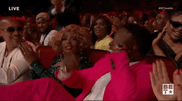 Celebrity gif. At BET Awards, Diddy reclines in his seat and kicks his legs and rolls his hands over each other, celebrating while others around him also clap and cheer.
