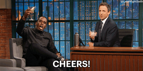 Late night gif. Seth Meyers and Sean Combs raise their glasses up while looking at us, both yelling, “Cheers!”