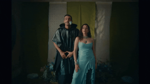 Music video gif. Jonita Gandhi and Ali Sethi in their music video for Love Like This. They're standing in the middle of a room and both simultaneously clap their hands twice to the beat. Jonita is wearing a shiny blue dress and Ali holds a rose in his mouth while he wears a black poncho that resembles a trash bag.