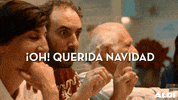 Video gif. A man with brown hair and a beard, sits near a woman. He raises one finger on each hand, then hits his hands together, revealing two fingers raised on one hand and closed fist on the other. He raises his eyebrows at his magic trick. Text, in Spanish "¡Oh! Querida Navidad."