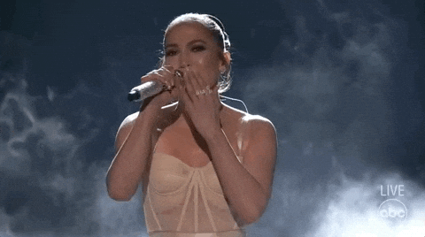 Celebrity gif. On a smoky stage, Jennifer Lopez smiles, blows a kiss to the audience and waves, and bows graciously.