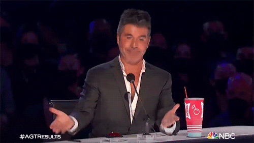 Reality TV gif. On the set of AGT, wearing a black jacket with a half-buttoned white shirt, Simon Cowell turns up his hands in a shrug without looking at us.