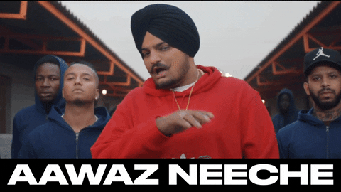 Video gif. Wearing a black turban and red hoodie, singer Sidhu Moose Wala holds his hand under his chin and waves it. A group of tough-looking men in blue hoodies stands behind him. Text, “Aawaaz Neeche.”