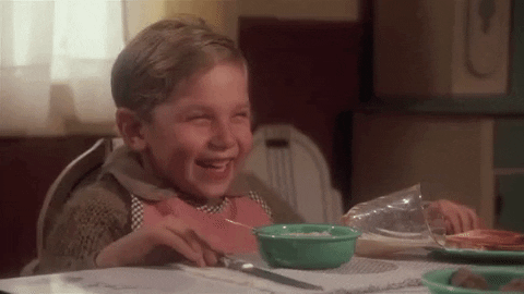 Movie gif. Ian Petrella as Randy in A Christmas Story sits at the dining table with a bib on as he laughs uncontrollably, leaning forward with his hands on the table, and then leans away. 