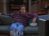Seinfeld gif. Jerry reclining on the couch with his feet up, eating popcorn and watching TV, making a victory fist in the air