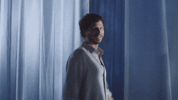 Sponsored gif. Michael Cera is wearing a silky, flowy light blue button up and he has a serene smile on his face while he spins through a room filled with the same colored soft fabric that drapes down from the ceiling. 