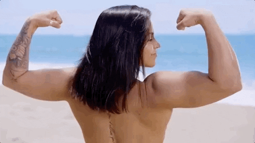 Reality TV gif. Muscular brunette woman from Ex on the Beach stands facing the ocean and flexes her biceps, looking over her shoulder and smiling at us.