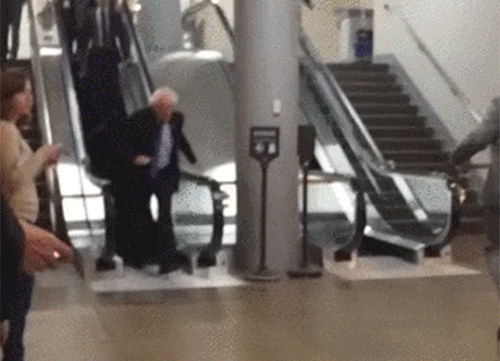 Political gif. We follow Bernie Sanders as he steps off of an escalator and jogs past several people.