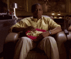 TV gif. Dule Hill as Gus on Psych sits with a bowl of popcorn on his lap. He picks up a piece of popcorn and bops his head around in happiness as he chews.