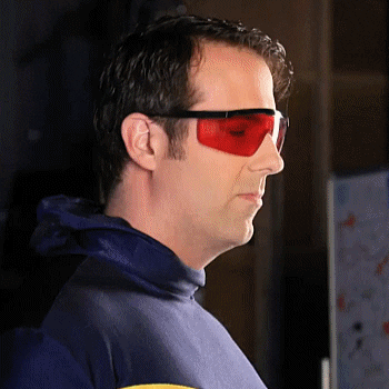 Video gif. Camera zooms in on a man wearing red lens sunglasses as he looks at us, winks, and a shimmer appears at his teeth.