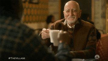 TV gif. Dominic Chianese as Enzo in The Village. He sits at a table and clinks mugs with someone with a satisfied smile.