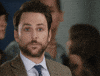 Movie gif. Wearing a brown suit jacket, Charlie Day gives us a blank look as several people walk by him in the background. After a second, he raises a hand to give us a thumbs up, then walks out of frame.
