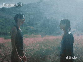 Sponsored gif. A man and woman stand facing each other as they gaze into each other's eyes, surrounded by a field of bright pink wildflowers in the pouring rain. A speech bubble appears near the woman that reads, "sup," and another one appears by the man that reads, "sup." The Tinder logo is in the bottom right corner.