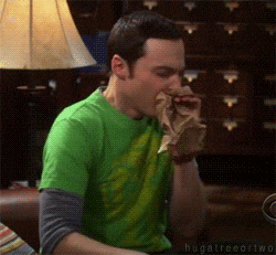 TV gif. Jim Parsons as Sheldon on The Big Bang Theory sits on a couch, breathing heavily into a paper bag.