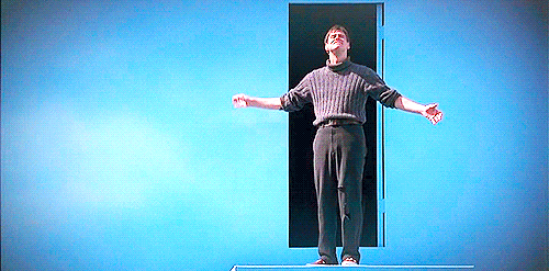 Movie gif. Jim Carrey as Truman in The Truman Show looks smug as he spreads his arms wide and takes a bow in front of a doorway in a sky blue wall.