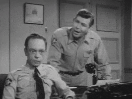 TV gif. In a scene from the Andy Griffith Show in black and white, Barney Fife as Don Knotts and Andy Griffith as Andy Taylor are dressed in officer uniforms and hover over a desk. Don, who is seated, puts his hands to his head and ruffles his hair violently with a baffled look on his face while Andy looks on, laughing.