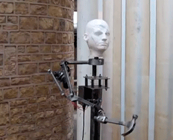Video gif. A disembodied robot arm with a plaster head, grasps a hammer and swings it awkwardly, smashing itself in the face.