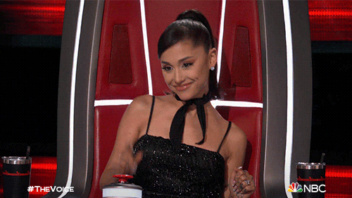 TV gif. Ariana Grande in a sparkling black dress smiles and points with pride from her judge's seat on The Voice. 