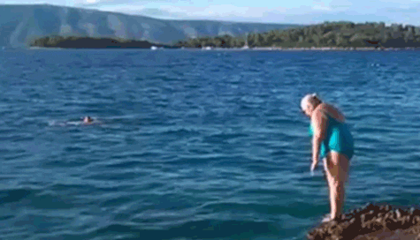 Video gif. Woman stands at the edge of a stony bank making a half-hearted attempt at a dive, bellyflopping into the water.