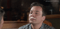 The Tonight Show gif. Jimmy Fallon tilts his head back and has a slice of sashimi on his lip like a white mustache. He reaches his tongue up and lets the fish go in his mouth. He then smirks and winks at the person across from him.